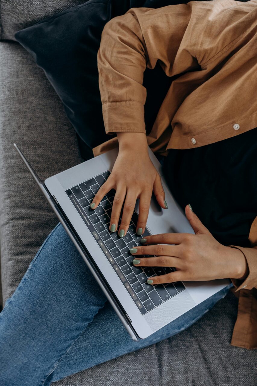 Person is sat on a sofa, wearing jeans and a brown shirt. An open laptop is resting on their lap., their fingers are on the keys.