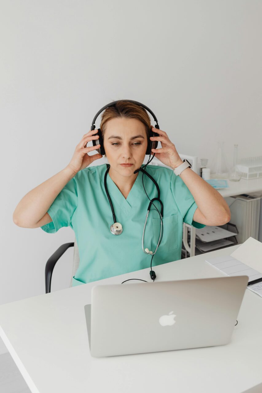 Medic wearing green scrubs is sat down in front of a computer with headphones on and a stethoscope around here neck.